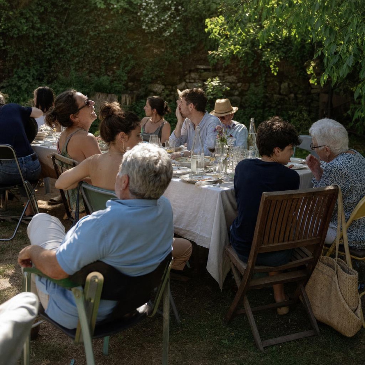 A long summer table in a garden at the end of a family meal, several generations in conversation, mismatched chairs and the remains of dessert, in natural afternoon light.