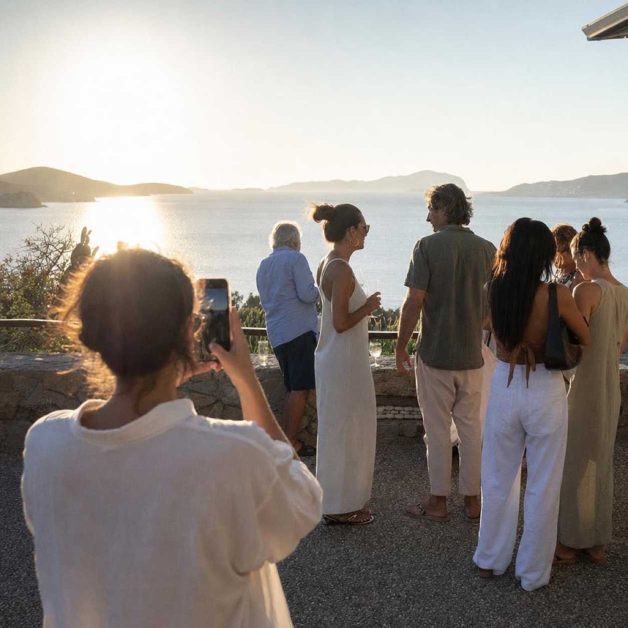 On a terrace above the sea at the end of a summer day, one person photographs a group of loved ones of all ages mid-conversation, glasses in hand, in the evening light.