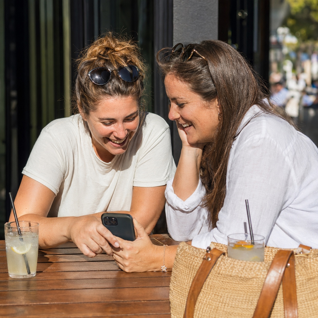 On a sunny summer terrace, two friends lean over a phone choosing holiday photos together, one laughing at a picture, two cold drinks on the table.