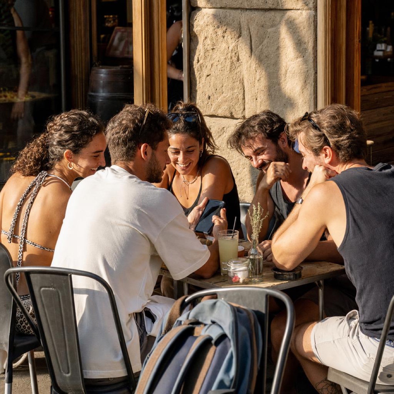 A group of friends at a terrace table at the end of the day, back from a summer trip, leaning over a phone, one of them laughing at a photo in the real evening light.